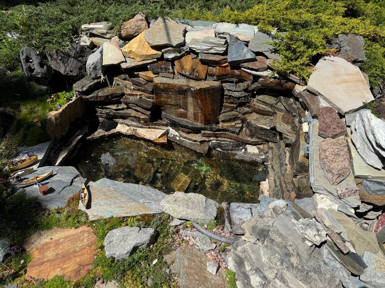 Miniature pond with clear water, tiny boats, and waterfall cascading over stacked rockwork in a garden railway