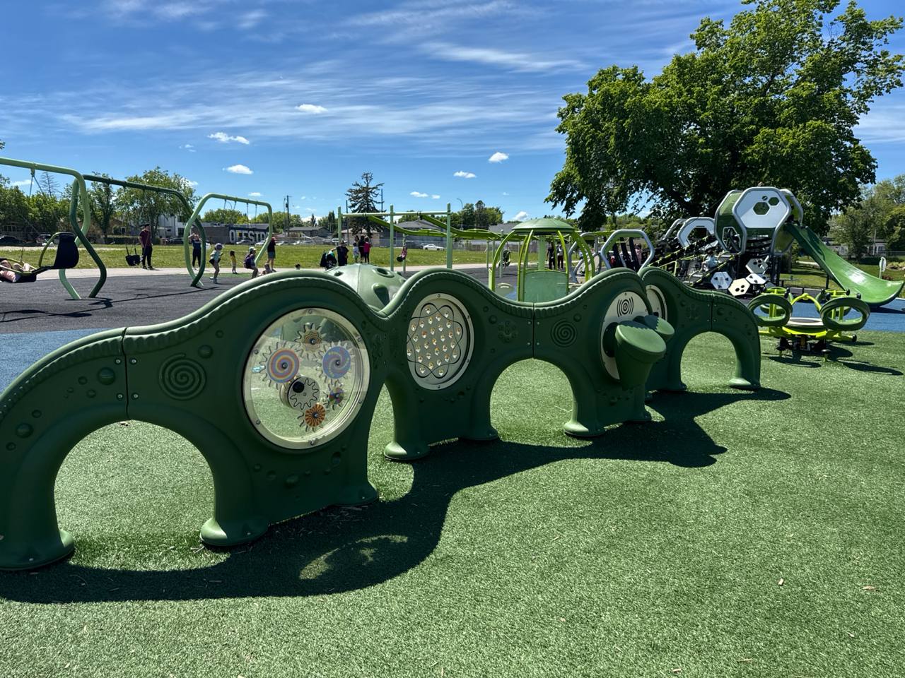 Sunny playground with green play structures, artificial turf, swings, and a slide under blue sky